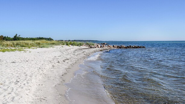 A sandy beach with clear water, a rocky breakwater, and distant trees.
