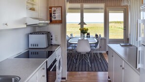 A compact kitchen with a toaster, a sink, and a washing machine. There is a dining area with a table and chairs, and a view of the beach through the door.