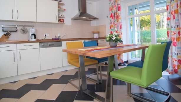 Eine moderne Küche mit weißen Schränken, einer Kaffeemaschine und einem Essbereich mit einem Holztisch und bunten Stühlen.