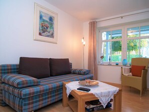 A living room with a plaid sofa, a wicker chair, a small table with a remote control, and a window with curtains.
