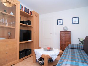 Un ambiente compatto con libreria in legno, televisione, tavolino con tovaglia di pizzo, divano e cassettiera.