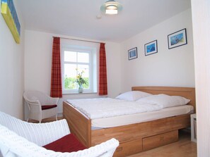 A bedroom with a wooden bed, white bedding, a wicker chair, a window with curtains, and three framed pictures on the wall.