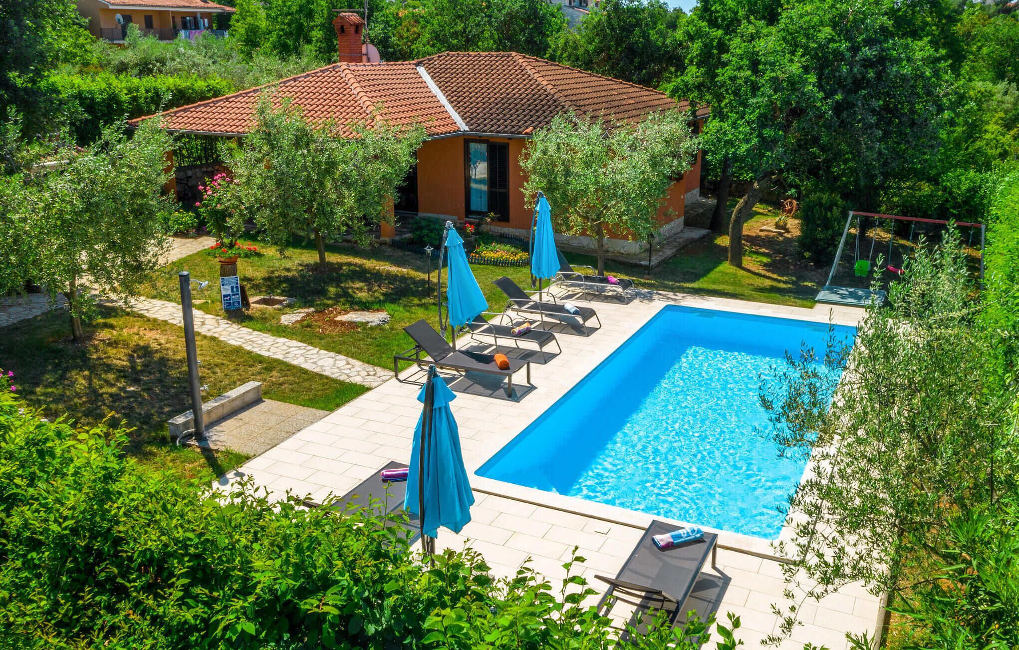 A rectangular swimming pool with lounge chairs and umbrellas, surrounded by lush greenery and a tiled deck.