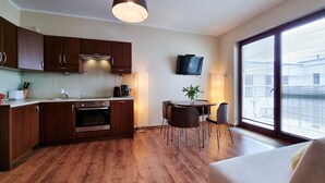 Een moderne keuken met houten kastjes, een inbouwoven en een eethoek met een tafel en stoelen. Een televisie is aan de muur bevestigd, en er is een raam dat zorgt voor natuurlijk licht.