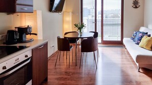 Een compacte keuken met een fornuis, koffiezetapparaat en een aan de muur bevestigde televisie. Er is een eethoek met een tafel en stoelen, plus een sofa met kussens.