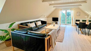 A spacious loft apartment with a black leather sofa, a dining table, and a large window offering a view of greenery.