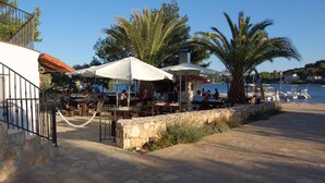 An outdoor dining area with tables and umbrellas, a stone wall, and a view of the sea and boats.