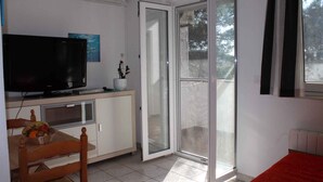 A room with a TV on a cabinet, a dining table with chairs, a sliding glass door to a balcony, and a red bed.
