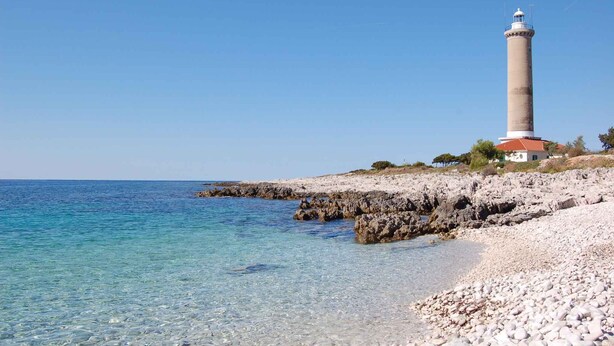 A coastal scene with a lighthouse on a rocky outcrop, clear blue water, and a pebble beach.