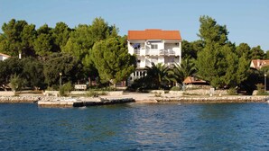 A two-story building with a red-tiled roof, surrounded by lush greenery and a waterfront.