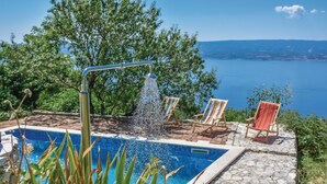 A pool with a water feature, surrounded by stone tiles and wooden chairs, overlooking a body of water and hills.