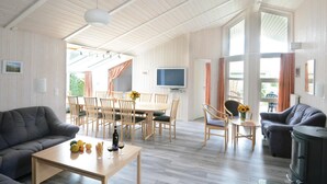 Een ruime woonkamer met houten plafond, een eettafel met stoelen, een bank, een aan de muur bevestigde televisie en een open haard.