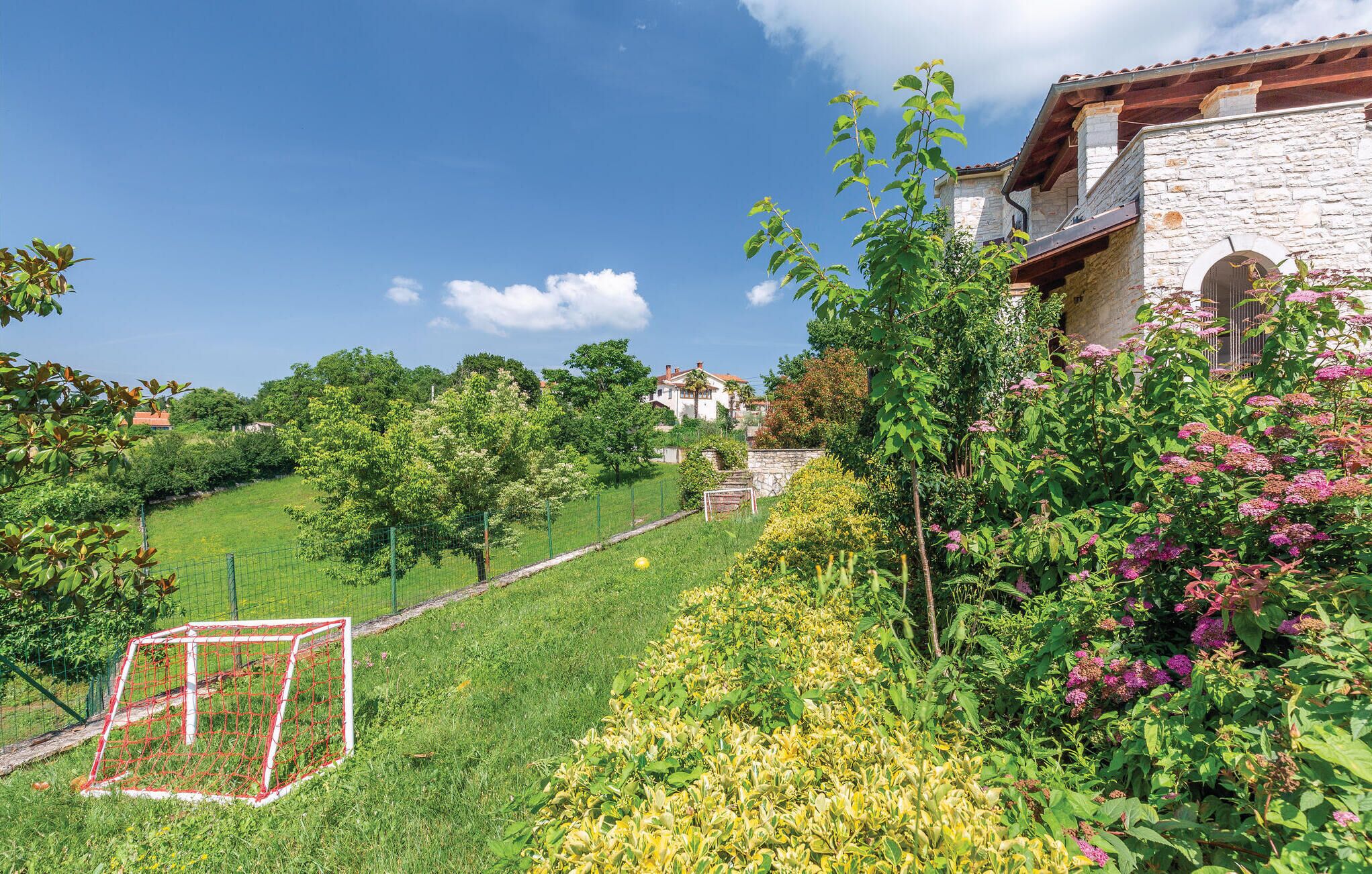 A lush garden with a soccer goal, a stone building with a red-tiled roof, and a clear blue sky.