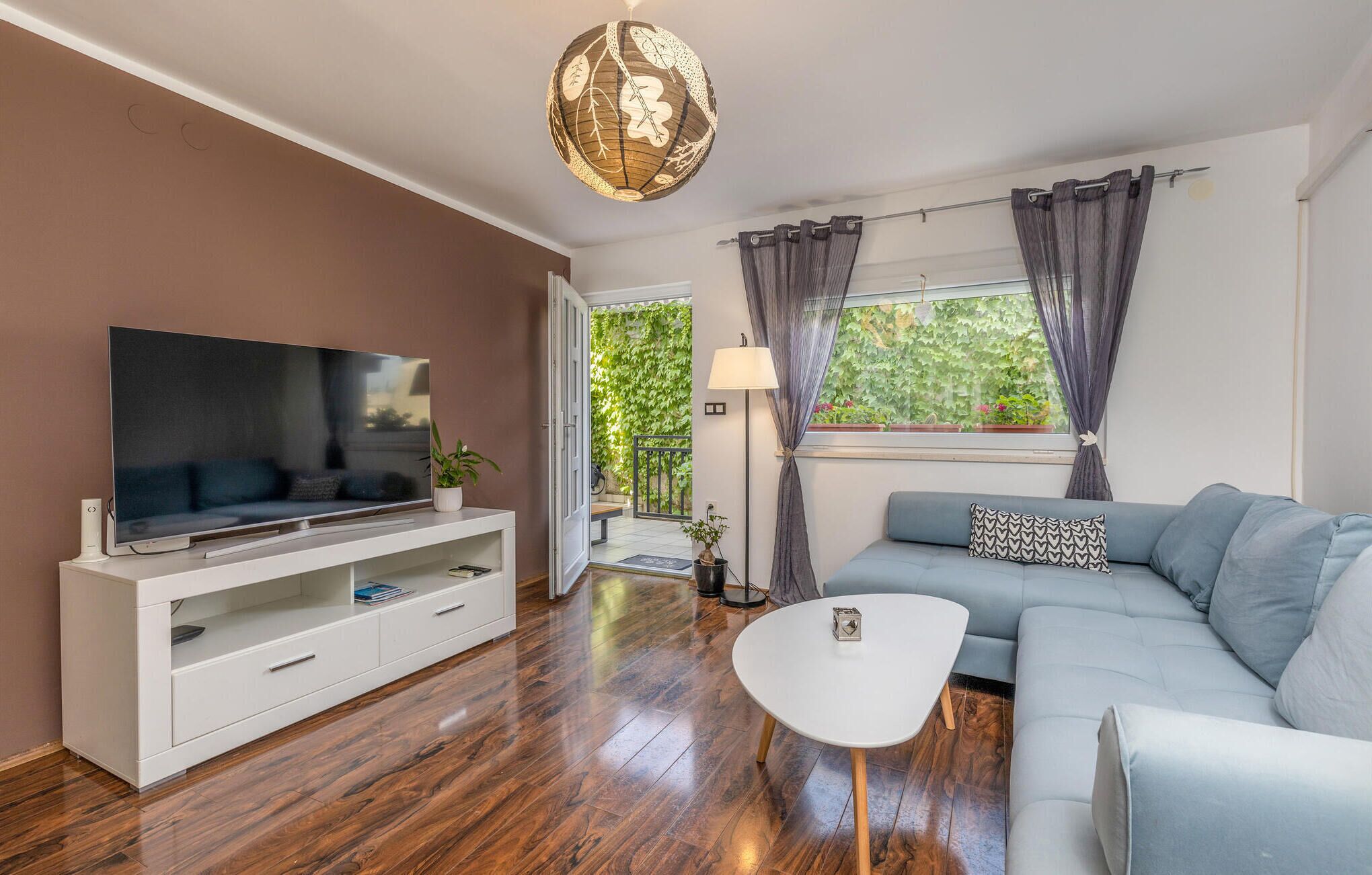 A modern living room with a flat-screen TV on a white console, a grey sofa with a patterned pillow, a white coffee table, and a large window with curtains.