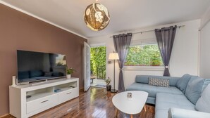 Un salon moderne avec un téléviseur à écran plat posé sur un meuble blanc, un canapé gris agrémenté d’un coussin à motifs, une table basse blanche et une grande fenêtre avec des rideaux.