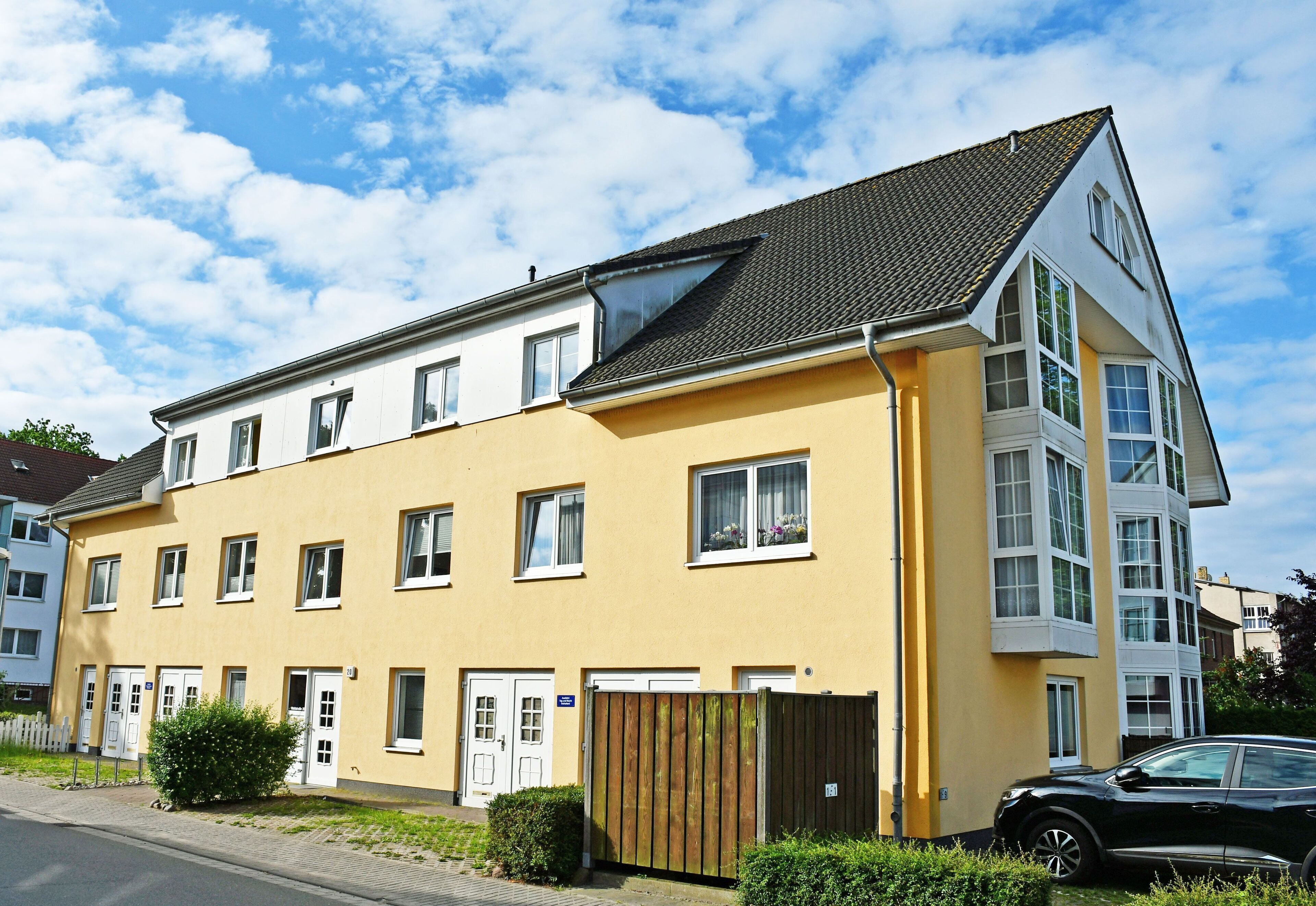 A two-story residential building with a yellow facade, multiple windows, and a black car parked in front.