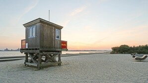 Una torretta di salvataggio in legno con un'antenna e due cartelli rossi, affacciata su una spiaggia sabbiosa con vista sullo skyline di una città in lontananza al tramonto.