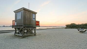 Ein hölzerner Rettungsschwimmturm mit Fahnenmast und zwei roten Schildern an der Seite, gelegen an einem sandigen Strand mit Blick auf eine entfernte Stadtlandschaft bei Sonnenuntergang.