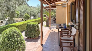 Un patio techado al aire libre con una mesa y sillas, rodeado de setos bien cuidados y una palmera.