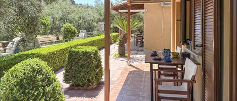 A covered outdoor patio with a table and chairs, surrounded by neatly trimmed hedges and a palm tree.