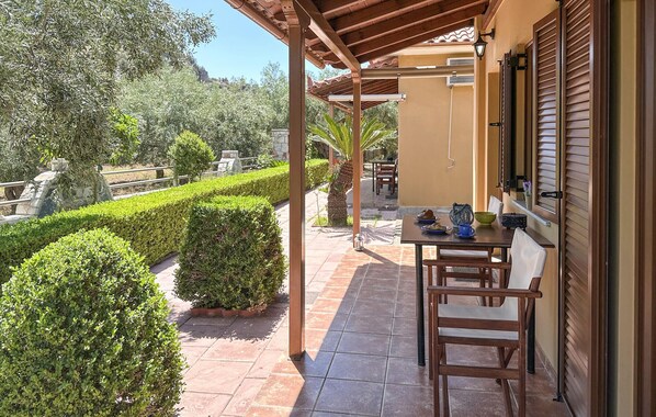 Un patio techado al aire libre con una mesa y sillas, rodeado de setos bien cuidados y una palmera.