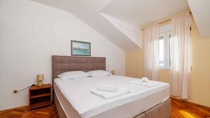 A neatly made bed in a room with wooden flooring, a nightstand with a lamp, and a window with curtains.