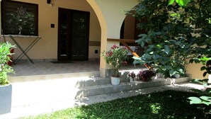 A building with a beige facade, a black door, and a small garden area with potted plants and a grassy foreground. - Holiday home with terrace and barbecue (Balatonboglár)