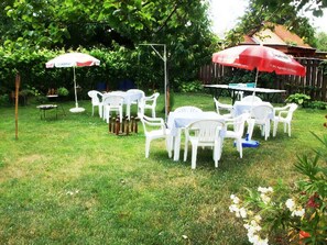 An outdoor seating area with white plastic chairs and tables, surrounded by greenery and a wooden fence.