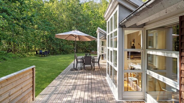 A wooden deck with a table and chairs, an umbrella, and a glass-enclosed room with a view of the greenery. - Nice home in Hasle with sauna (Hasle)