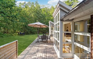 Une terrasse en bois avec une table et des chaises, un parasol et une pièce fermée par des baies vitrées, offrant une vue sur la verdure.