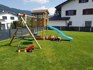 Une aire de jeux avec un toboggan et des structures d’escalade, entourée d’une zone délimitée par une clôture et dotée d’une pelouse.