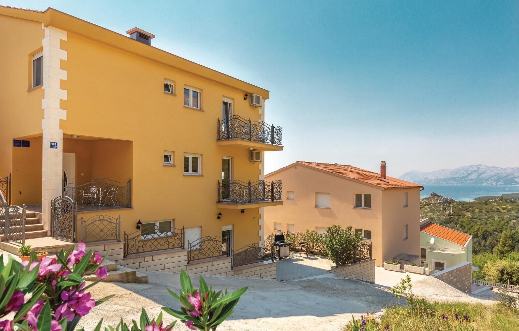 A yellow multi-story building with balconies, a small attached structure, and a view of the sea and mountains.