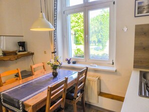 A dining area with a table, chairs, a vase with flowers, a hanging light, and a window with a view of greenery.