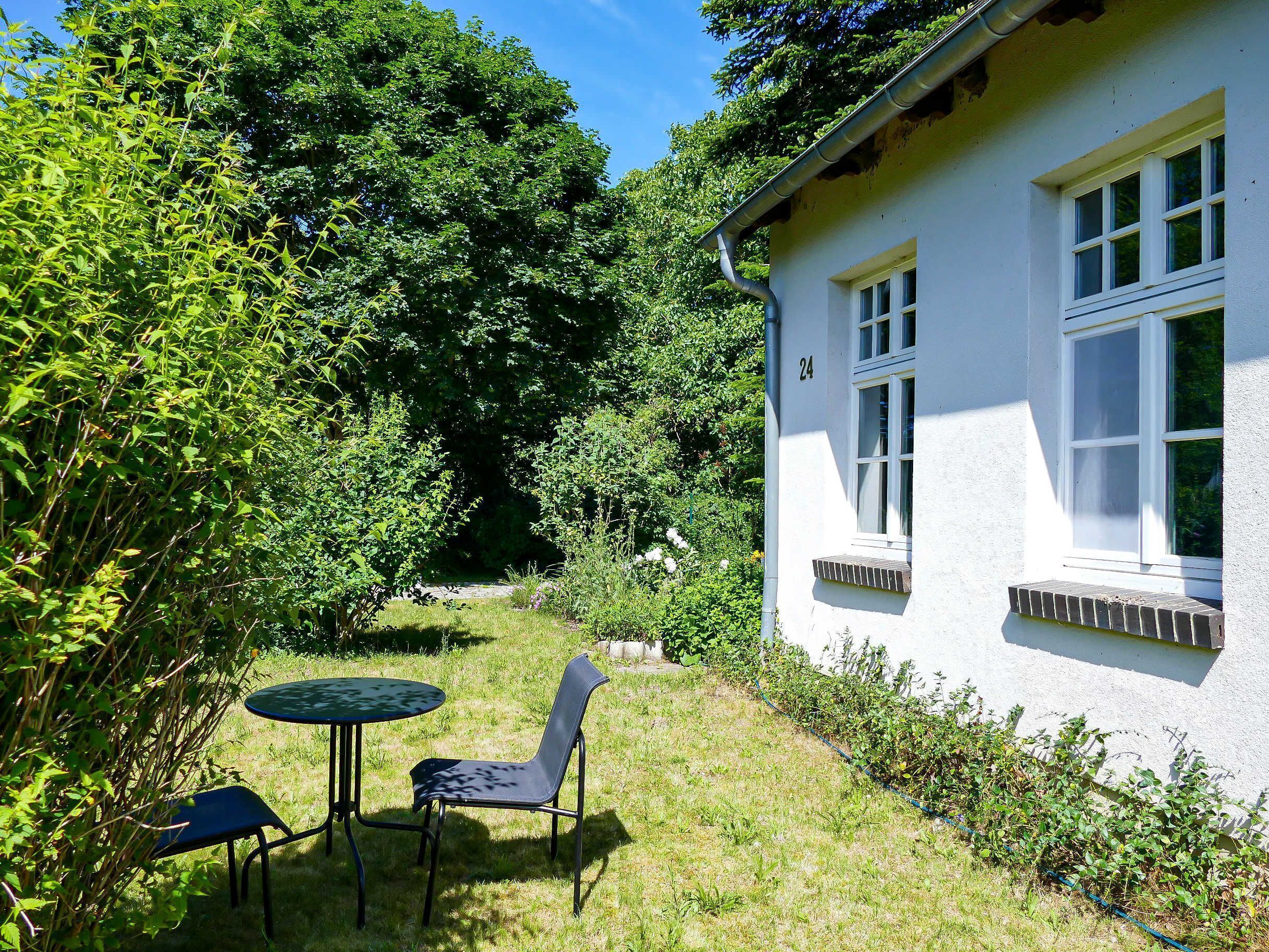 A white house with a number 24 on the wall, a small garden with a table and two chairs, and dense greenery surrounding the property.