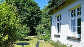 Ein weißes Haus mit der Nummer 24 an der Fassade, ein kleiner Garten mit Tisch und zwei Stühlen, umgeben von üppiger Vegetation.