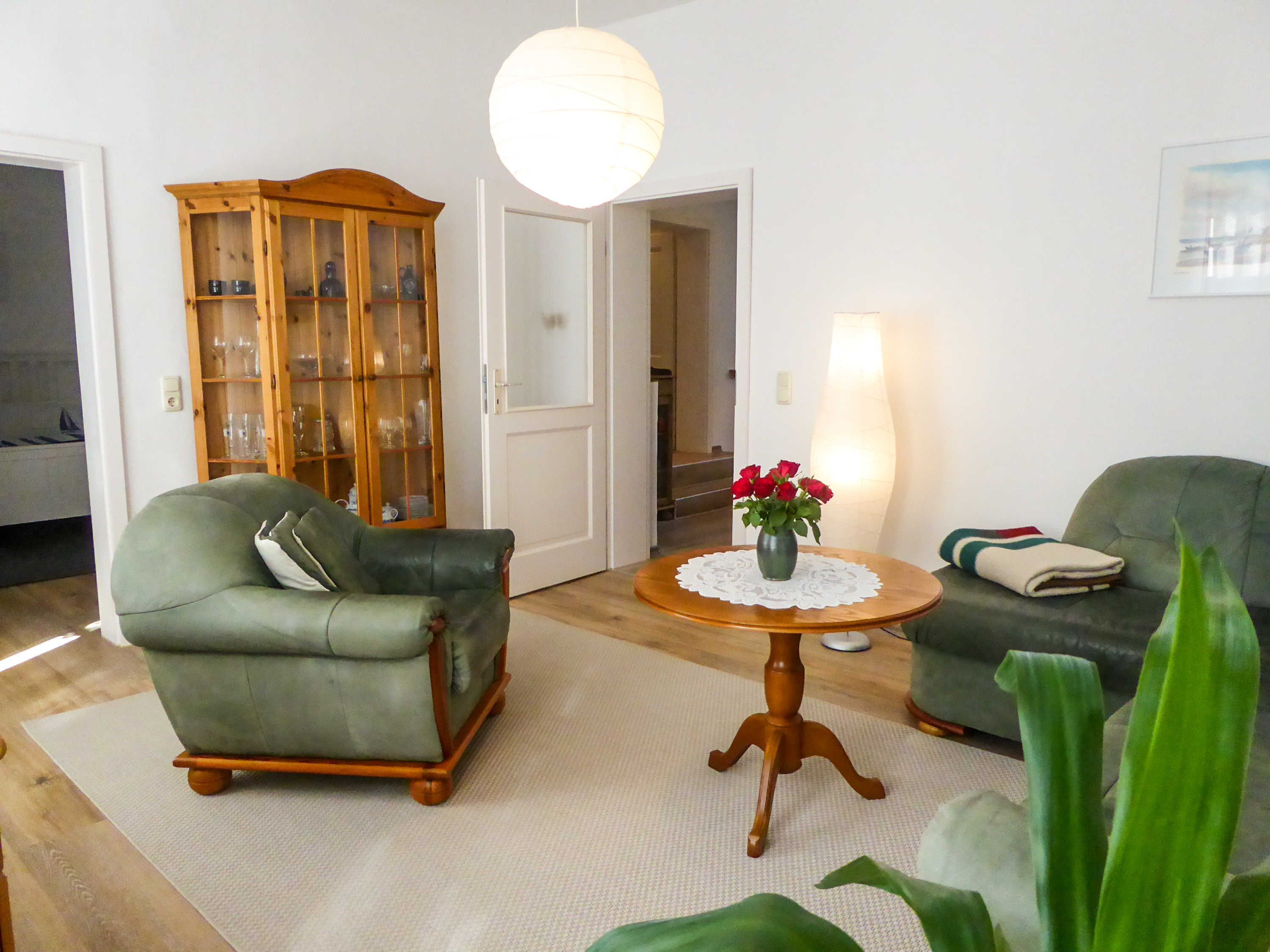 A living room with a green armchair, a wooden cabinet, a round table with a vase of flowers, and a lamp.