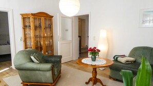 Ein Wohnzimmer mit einem grünen Sessel, einer Holzkommode, einem runden Tisch mit Blumenvase und einer Lampe.