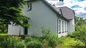 Ein kleines, eingeschossiges Gebäude mit geneigtem Dach, umgeben von üppigem Grün und blühenden Pflanzen.