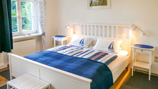 Ein Schlafzimmer mit einem Bett, zwei Nachttischen, einem Fenster mit Vorhängen, einem Heizkörper und einem Bild an der Wand.