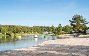 A lakeside view with a sandy beach, a row of cabins, and a forested area in the background. - Lovely apartment in Faßberg/Heidesee (Faßberg/Heidesee)