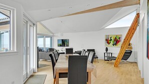 Ett rymligt matområde med ett långt träbord och svarta stolar, ett modernt kök i bakgrunden och ett stort fönster som släpper in naturligt ljus.
