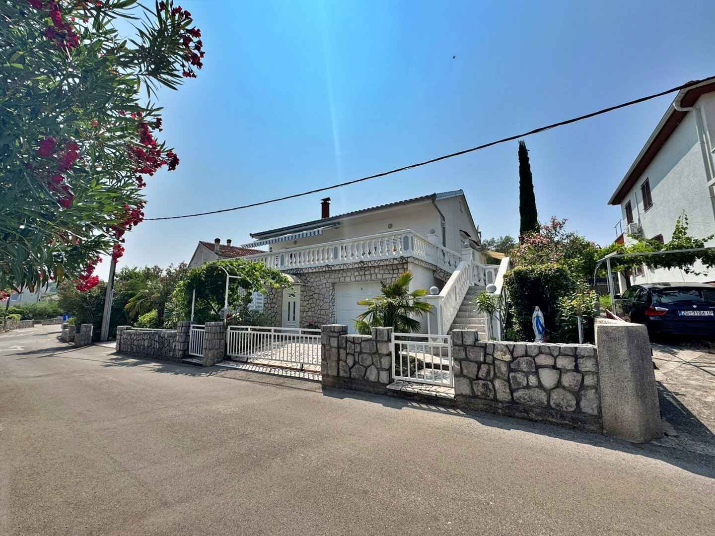 Ein zweistöckiges Haus mit einer Steinfassade, einem weißen Balkon und einer Steinmauer, die das Grundstück umgibt.