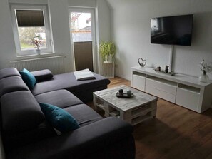 Uma sala de estar moderna com um sofá modular cinza, uma mesa de centro de madeira, um rack branco e uma televisão de tela plana fixada na parede.