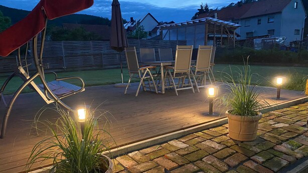 Una terraza de madera con mobiliario exterior, plantas en macetas y una pérgola con sombrillas.