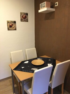 Uma área de refeições com uma mesa posta para quatro pessoas, cadeiras brancas, toalha de mesa preta e um prato de madeira, além de duas fotos penduradas na parede.