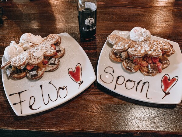 Dos platos de postre con la palabra "Fellow" y "Spoon" escritos, sobre una mesa de madera con una botella de Fritz-Kola al fondo.
