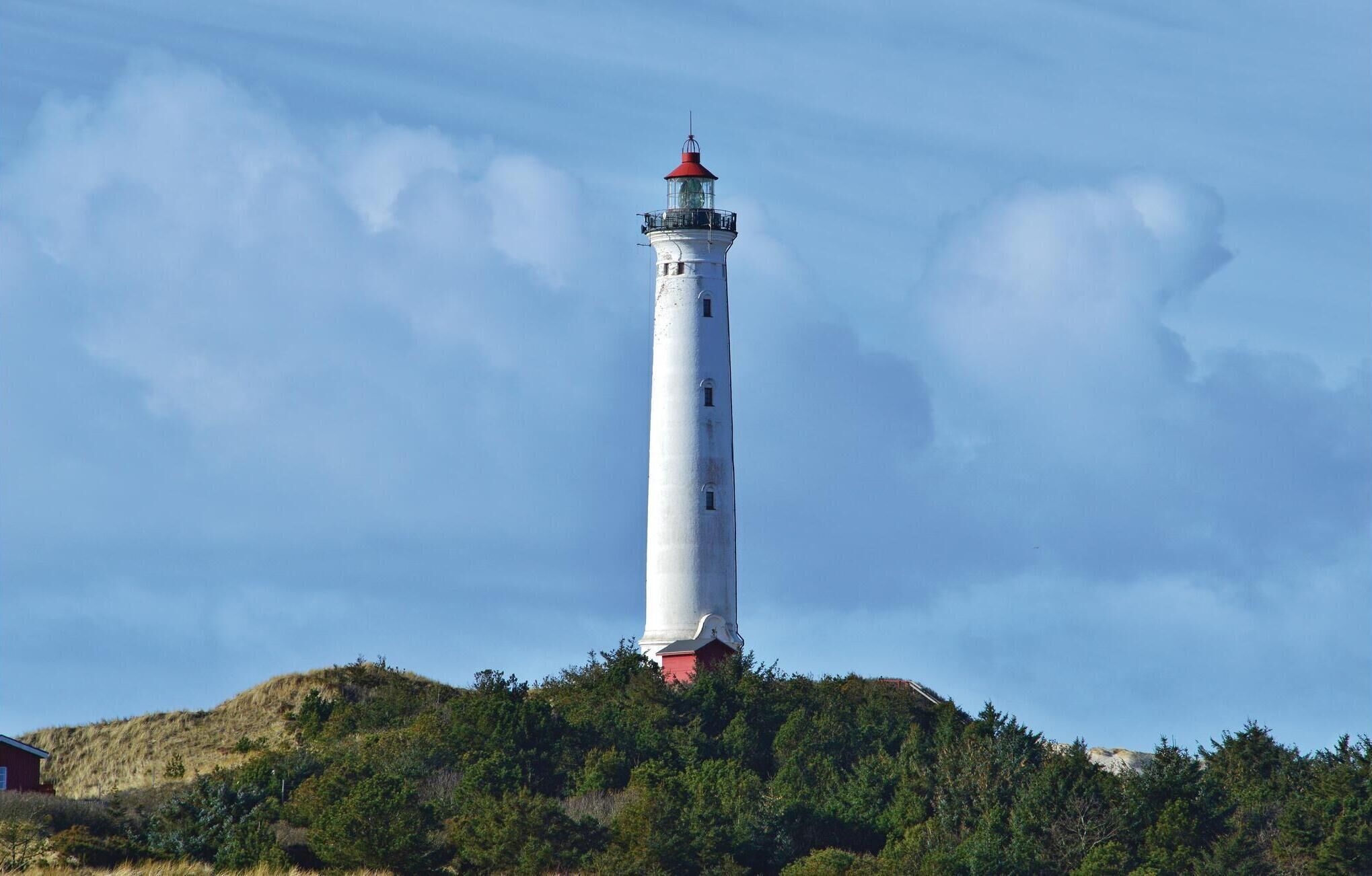 A tall white lighthouse with a red top, situated on a hill with dense green vegetation.