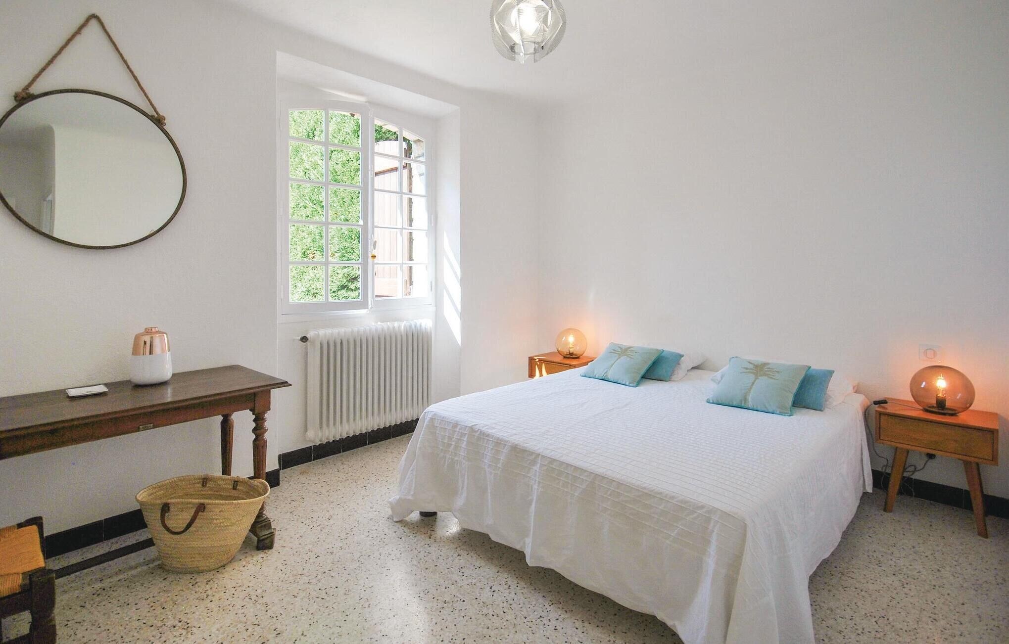 A bedroom with a bed, bedside lamps, a window, a radiator, a mirror, a wooden table, and a wicker basket.