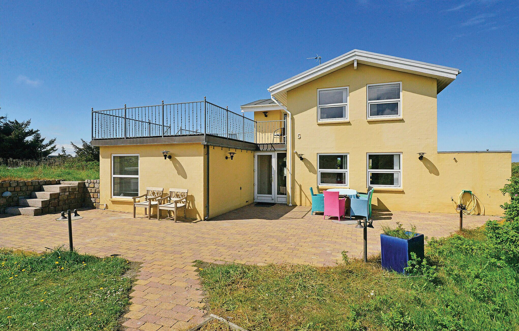 A two-story yellow house with a balcony, a patio with chairs, and a small garden area.