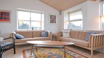 Un salon avec deux canapés, une table basse ronde et un tapis à motifs. Il y a deux fenêtres avec des stores, un tableau au mur et une lampe sur une table d’appoint.